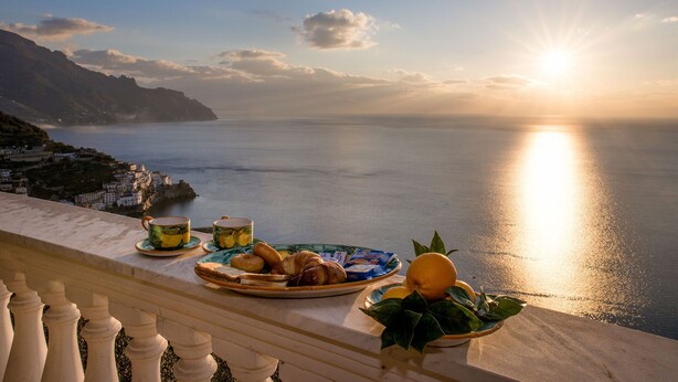 A balcony overlooking a coastal view with a table set for breakfast including croissants, a plate of pastries, a glass of juice, and a bowl of fruit. - Villa Gioiello (Amalfi)