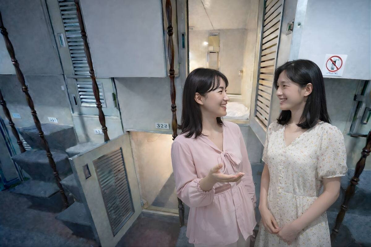 Two people standing in a narrow hallway with a staircase and a vent on the wall.