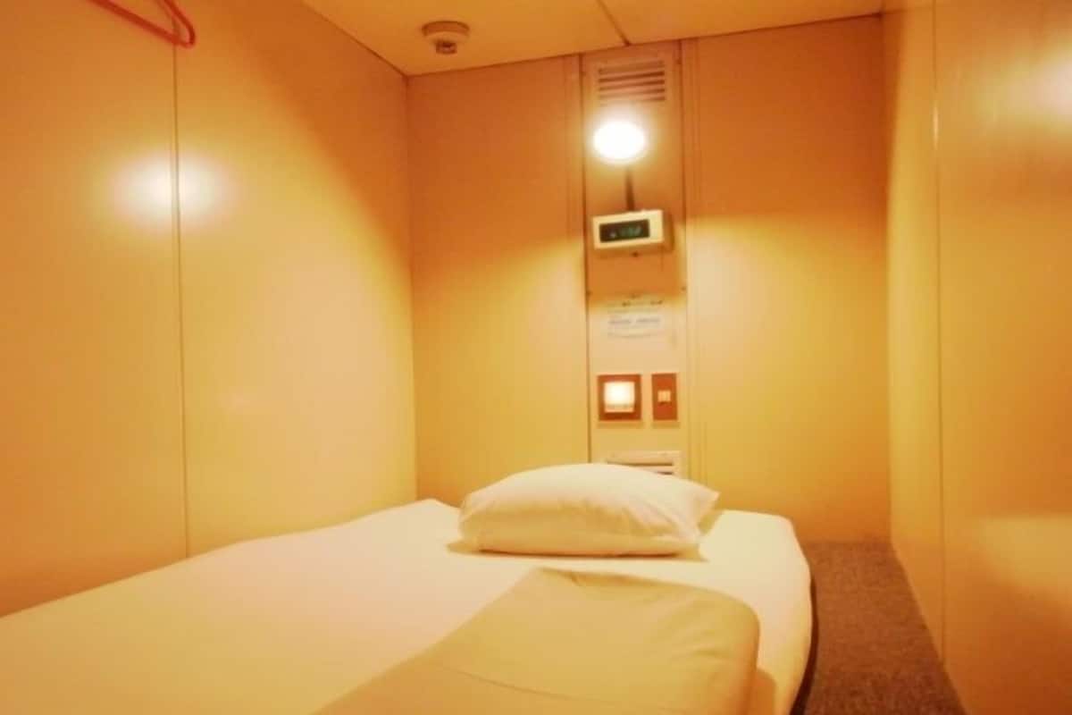A small, single-bed room with a bed, a wall-mounted control panel, and a door.