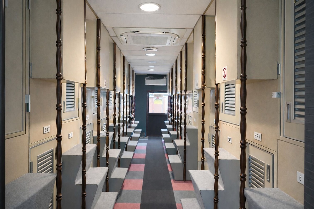 A hotel corridor with lockers on both sides, numbered room signs, and a checkered carpet.