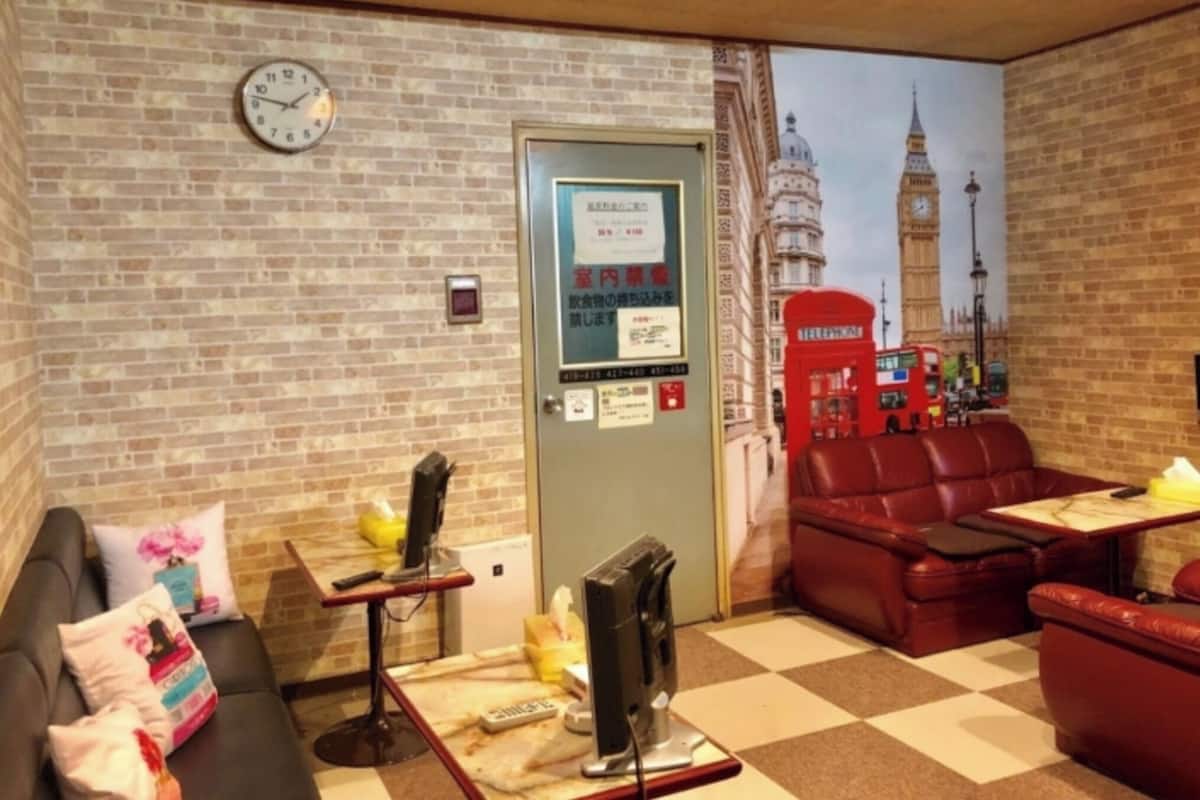 Una stanza con parete in mattoni, un orologio, una porta con un poster, un divano rosso, un tavolino con un computer e un pavimento a scacchi.