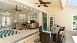A spacious living area with a glass dining table, wicker chairs, a ceiling fan, and a large window. - Sunside House, 3/2 Pool Home in Gated Community and minutes from everything. (Naples)