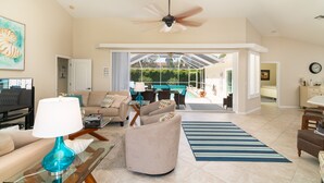 A spacious living room with a television, comfortable seating, a ceiling fan, and a view to an outdoor area. - Sunside House, 3/2 Pool Home in Gated Community and minutes from everything. (Naples)