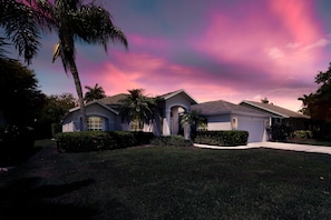 A two-story house with a garage, surrounded by well-maintained lawns and palm trees. - Sunside House, 3/2 Pool Home in Gated Community and minutes from everything. (Naples)