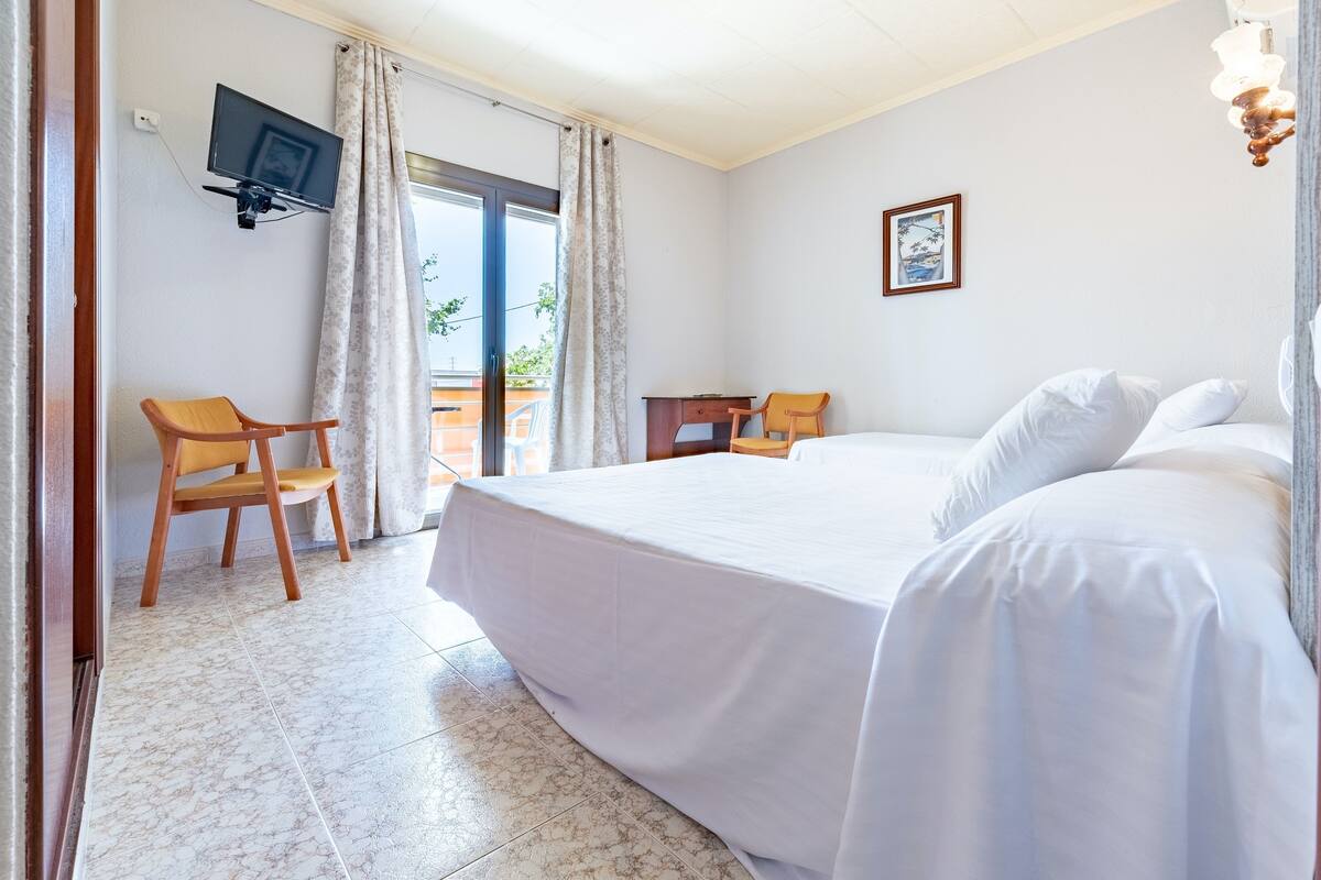 A hotel room with a large bed, a desk, a chair, a TV mounted on the wall, and a balcony with curtains.