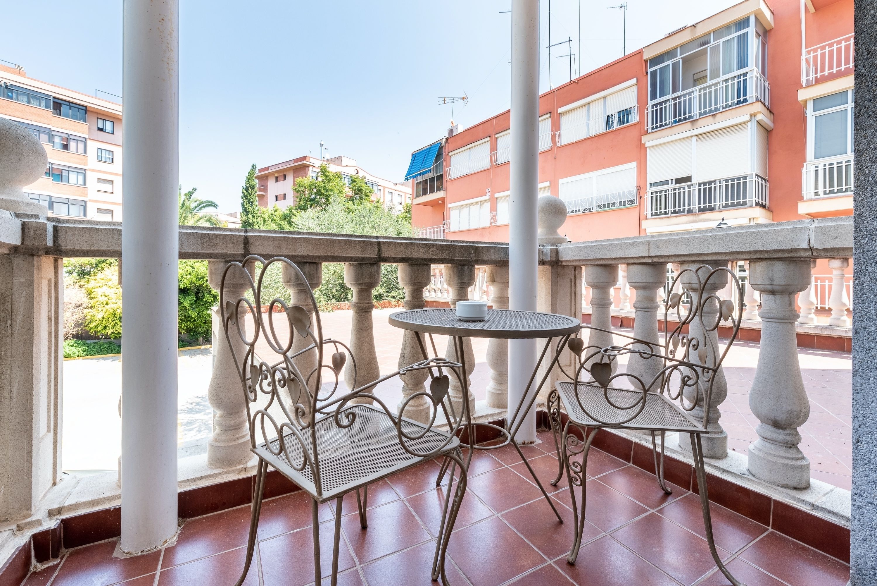 A balcony with two metal chairs and a small round table, overlooking a residential area with multi-story buildings.
