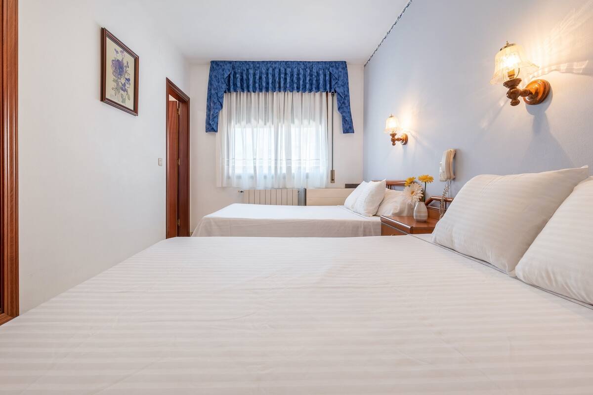A hotel room with two beds, a window with a blue valance, a bedside table with a lamp, and a wall-mounted phone.