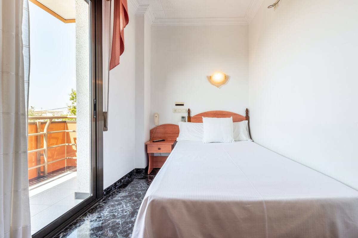 A hotel room with a bed, a bedside table, a wall-mounted light, and a view of the outside balcony.