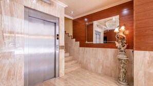 A modern elevator with a metallic finish, a marble floor, and a wooden wall with a mirror and decorative light fixture. - Hotel Canadá (Tarragona)