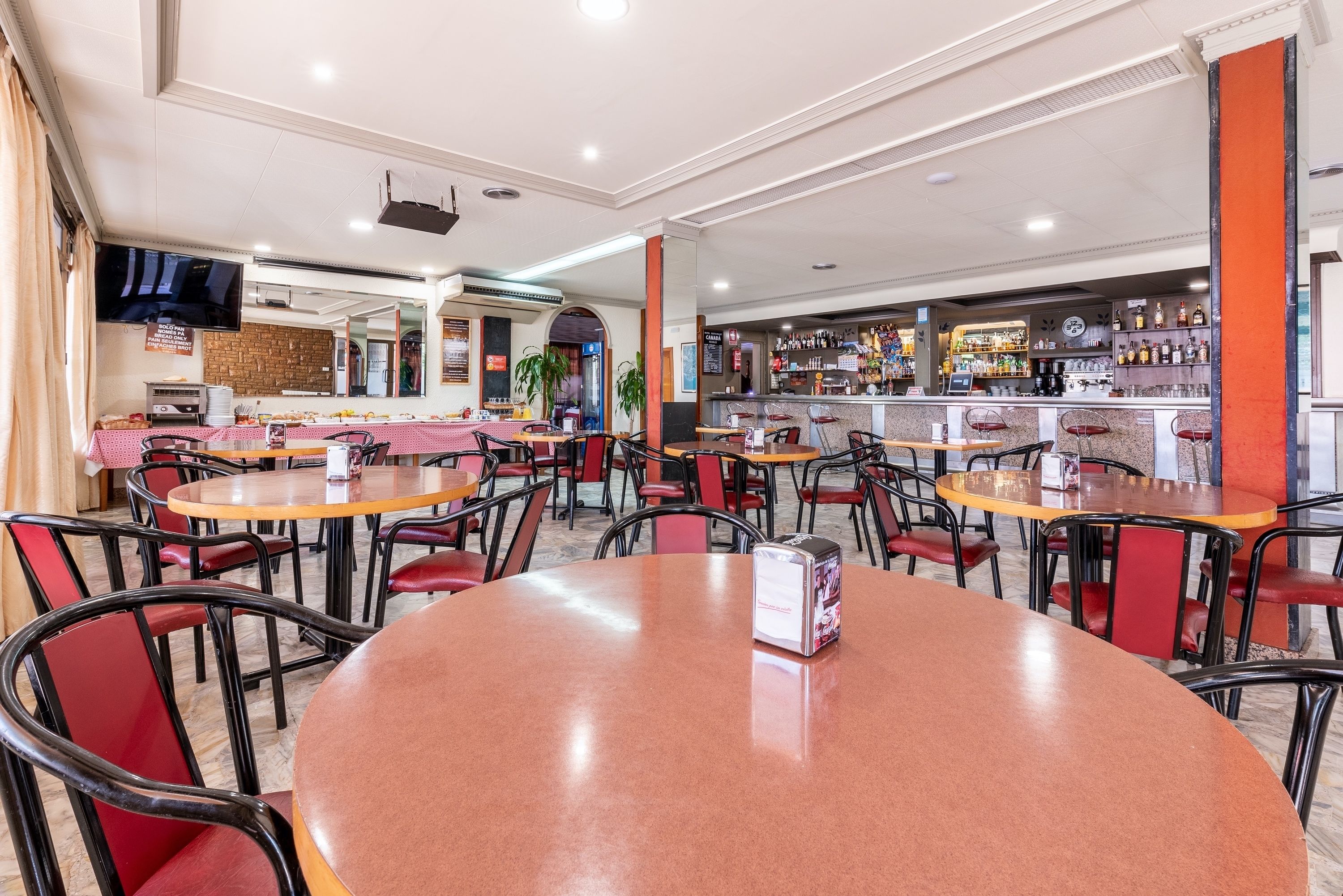 Een restaurant met ronde tafels en rode stoelen, een bar met flessenplanken, en een aan de muur bevestigde televisie.
