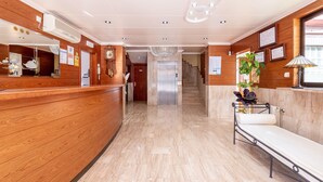 A reception area with a wooden reception desk, a waiting bench, a chandelier, and a staircase leading to an elevator. - Hotel Canadá (Tarragona)