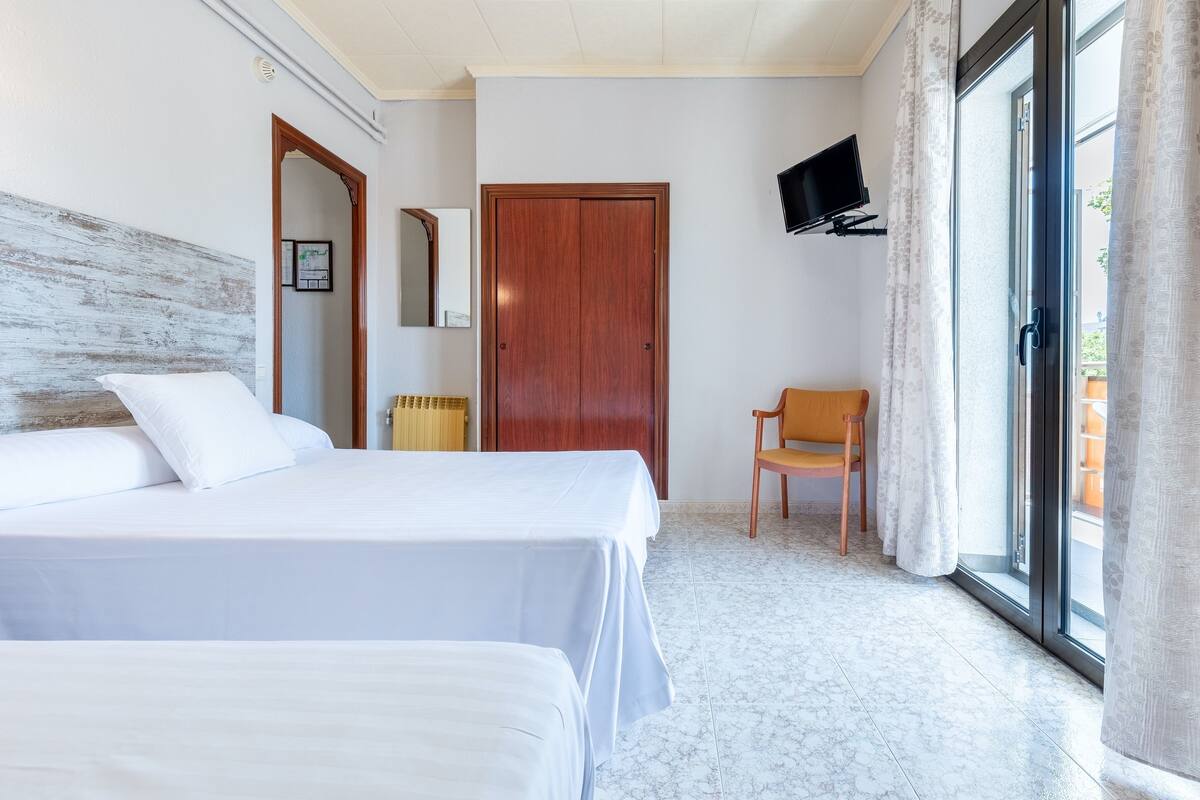 A hotel room with two beds, a wooden door, a TV mounted on the wall, and a chair by the window.