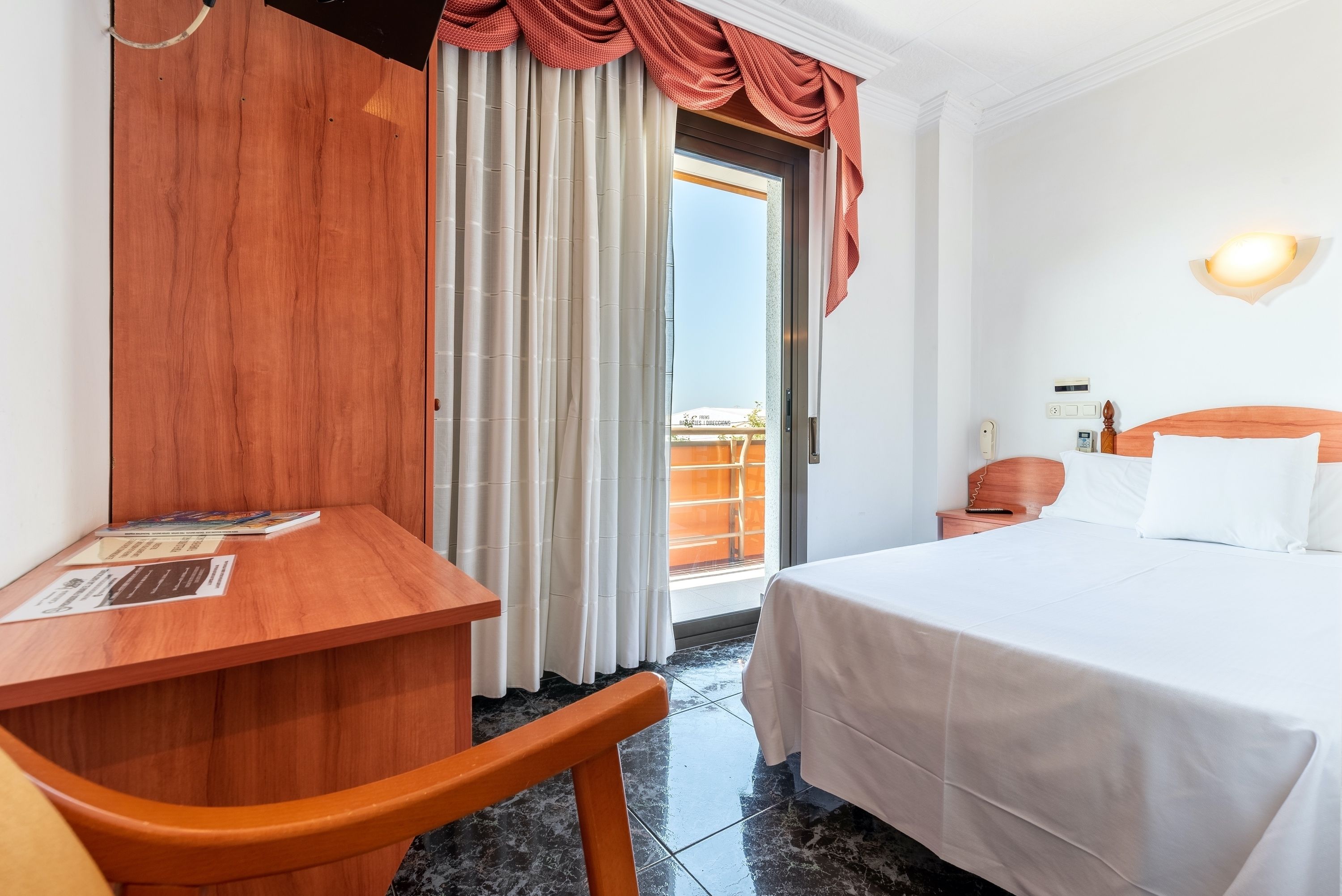 A hotel room with a bed, a desk, a chair, and a view of the outside balcony.