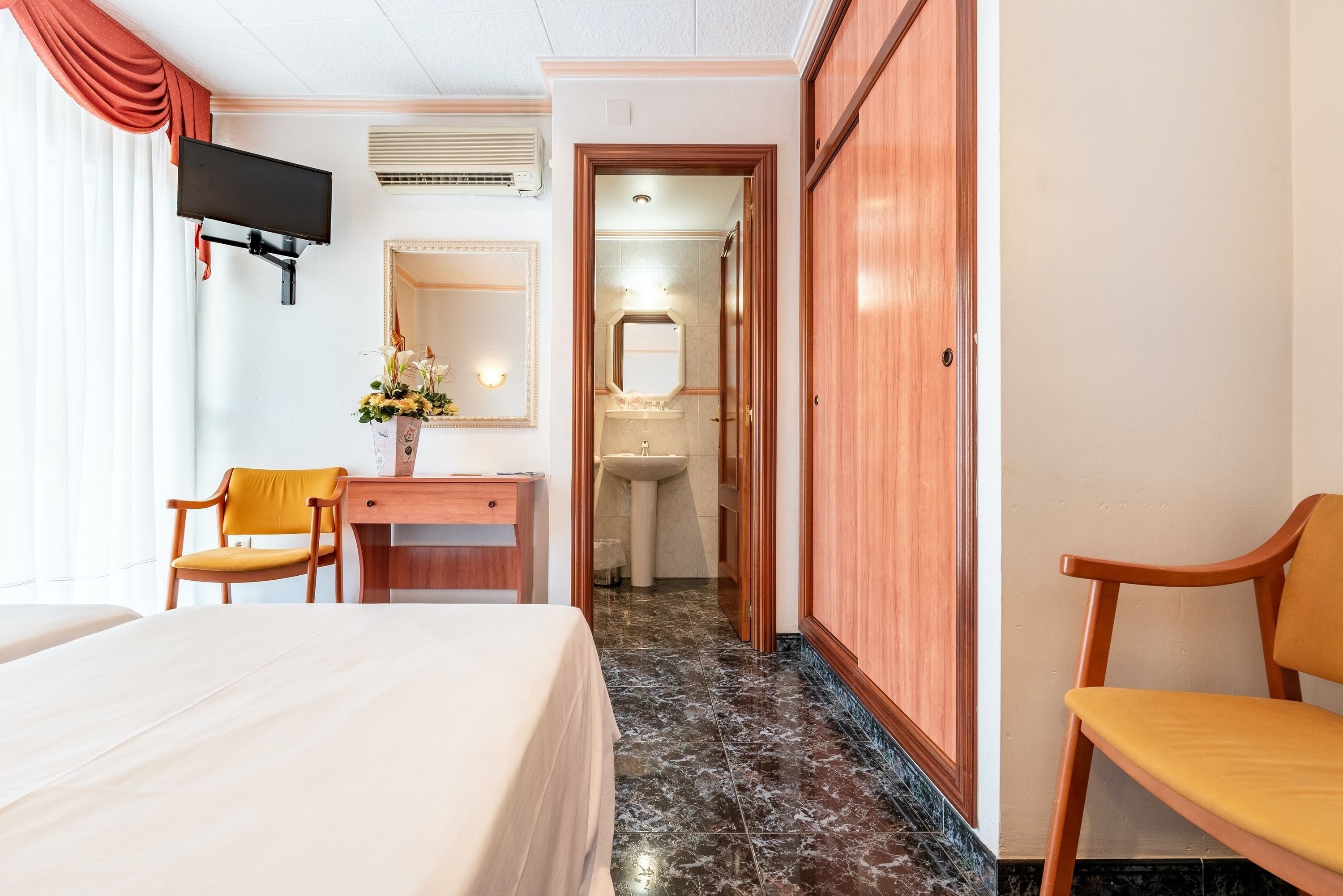 Habitación de hotel con cama, escritorio, silla, televisor, espejo y baño visible a través de una puerta abierta.