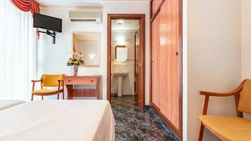 Habitación de hotel con cama, escritorio, silla, televisor, espejo y baño visible a través de una puerta abierta.