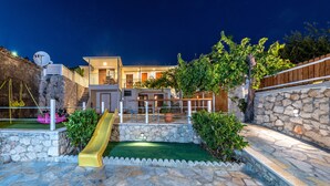 A modern house with a stone exterior, a yellow slide, and a well-lit patio area. - Stoned Traditional House just 3km from amazing beaches (Kalamitsi)