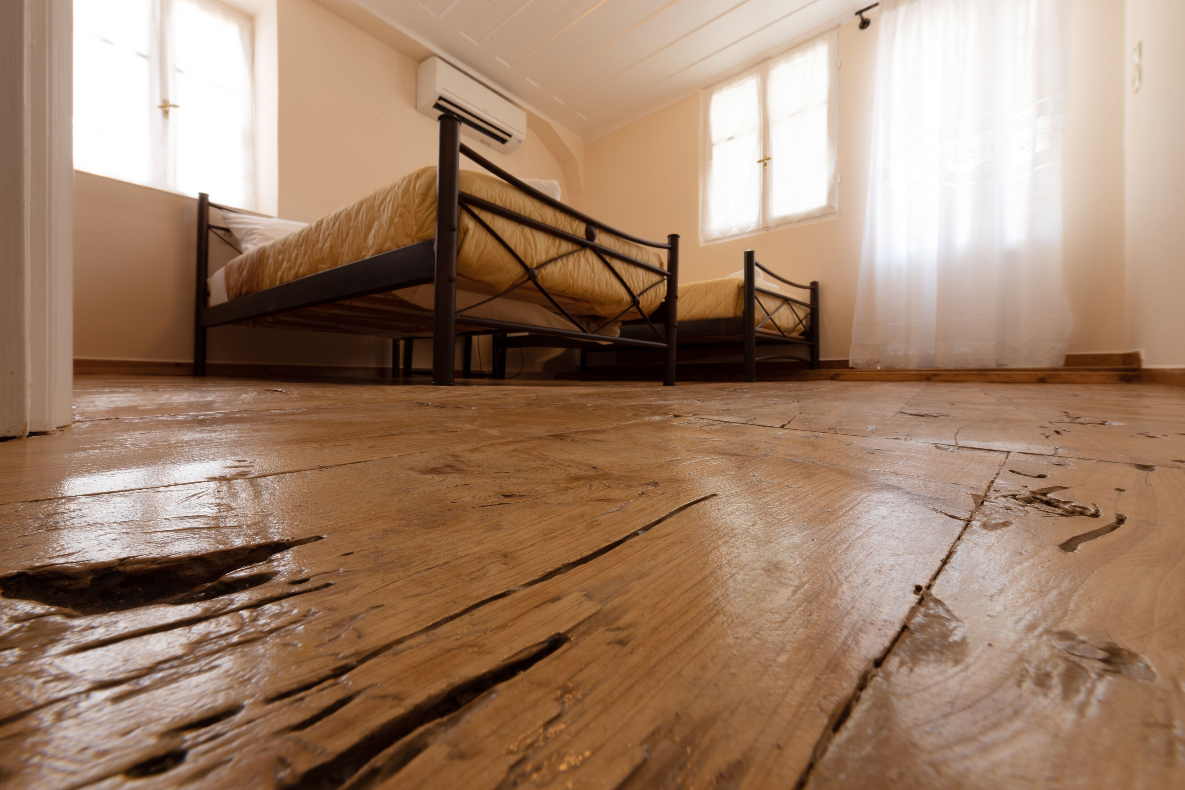 Een kamer met een houten vloer, een bed met metalen frame en een raam met dunne gordijnen.
