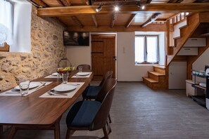 A dining area with a long wooden table set for four, stone walls, a wooden staircase, and a door leading to another room.