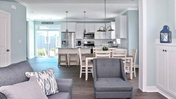 A modern kitchen with white cabinets, stainless steel appliances, and pendant lighting. There is a dining area with a wooden table and chairs, and a gray sofa with a patterned pillow. - Spacious Beach Retreat Near VB Boardwalk! (Virginia Beach)