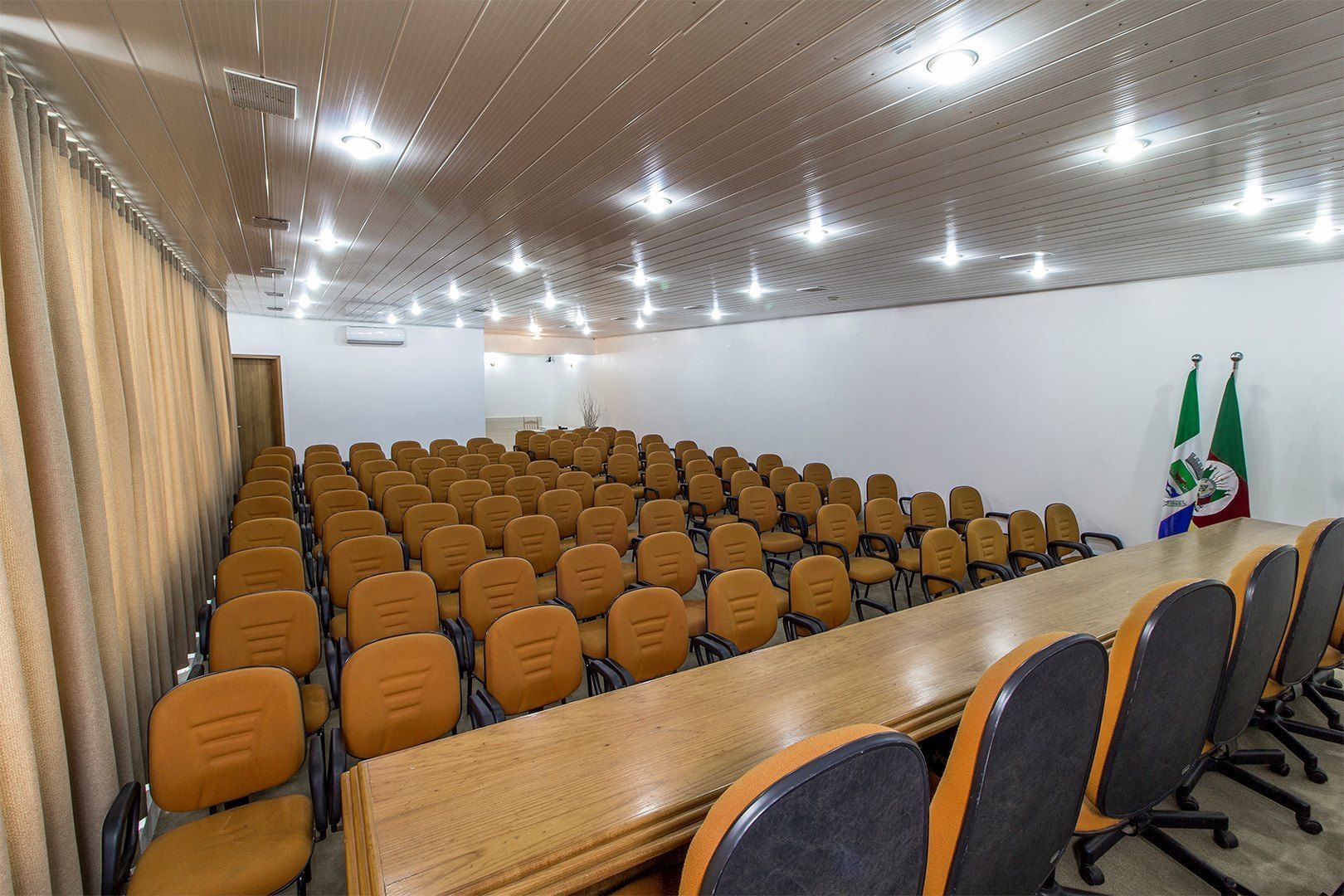 Un salón de conferencias con filas de sillas naranjas, una mesa de madera larga y dos banderas a la derecha.