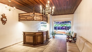 A reception area with a wooden reception desk, a chandelier, a sofa, and a view of a bus through glass doors. - Hotel Kimar (Tramandai)