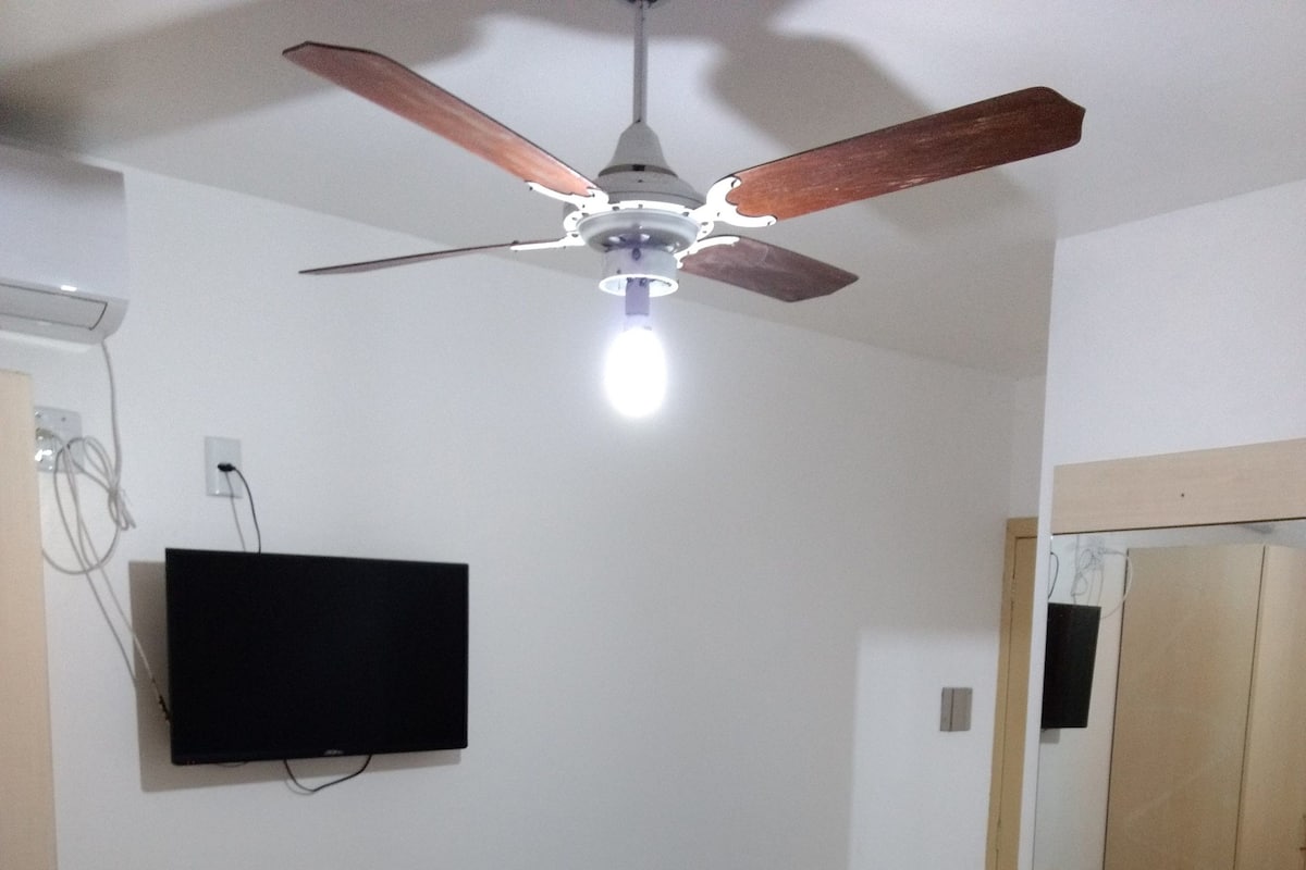 Ein Schlafzimmer mit einem Bett, einem Schreibtisch, einem Stuhl, einem Deckenventilator und einem an der Wand montierten Fernseher.