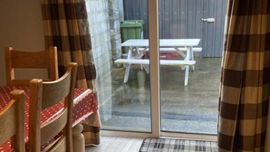 Ein Zimmer mit einem Holzbett, einem Stuhl und einem Tisch, bedeckt mit einer roten Tischdecke. Draußen steht ein Picknicktisch und eine grüne Mülltonne.