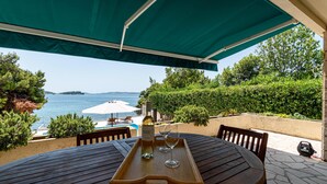 A wooden table with a tray, a bottle of Malvasia Starska wine, and two glasses, set under a green canopy overlooking a body of water and a mountainous landscape.