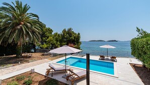 A pool area with a view of the sea, surrounded by palm trees and a pergola with umbrellas.