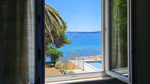 A view from a window overlooking a swimming pool with a clear blue sea and a distant shoreline.