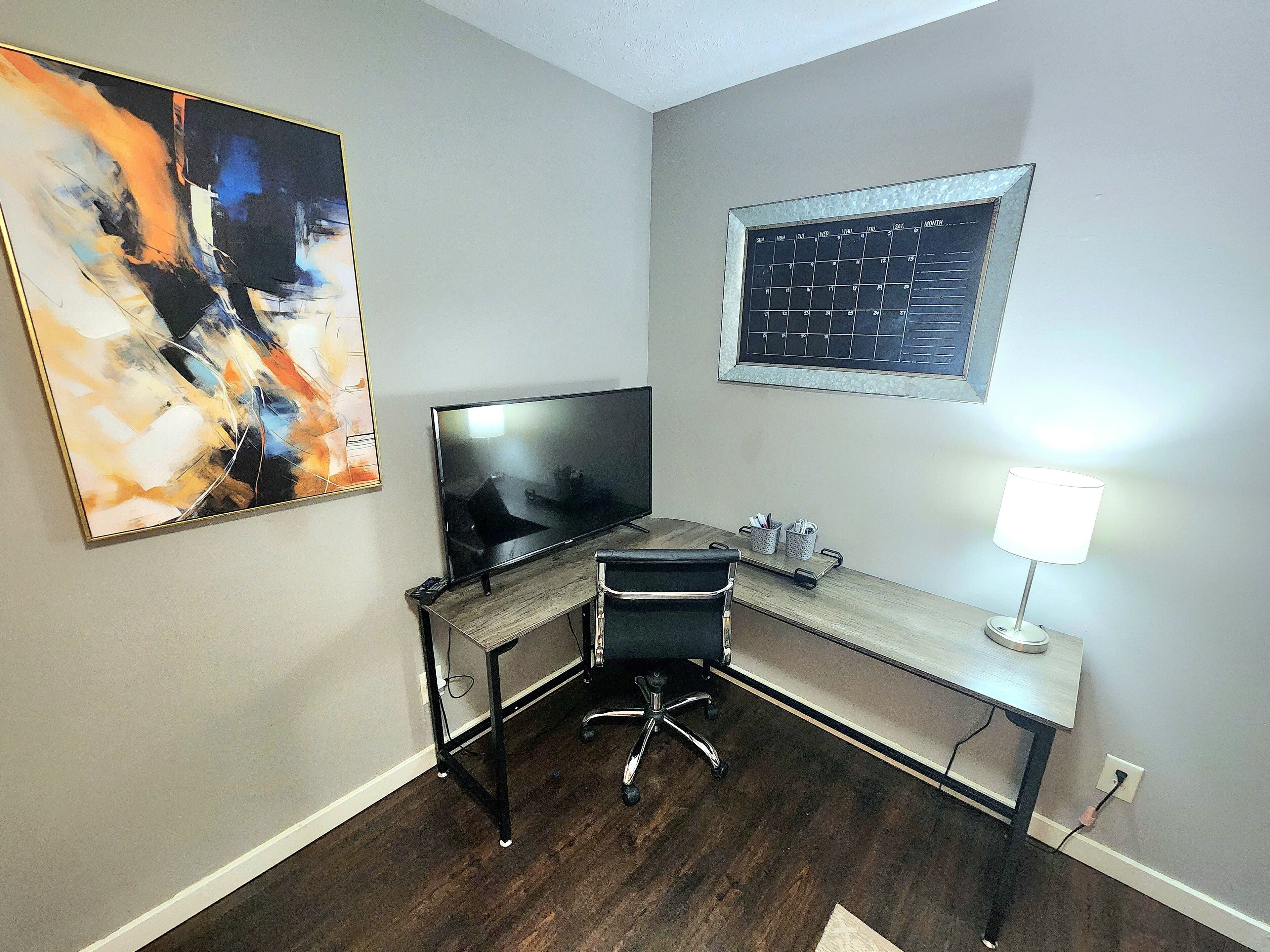 A modern office space with a desk, chair, TV, abstract painting, calendar, and lamp.