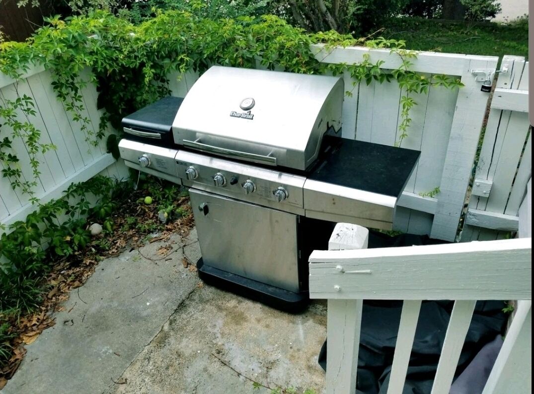 A stainless steel barbecue grill with a lid, a side burner, and a drawer, set against a white wooden fence with greenery.