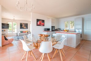 A spacious living area with a glass dining table, white chairs, a sofa, and a TV.