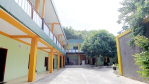 A spacious courtyard with a covered walkway, a green tree, and a building with yellow and green walls. - Losmen Alden (Parangtritis)