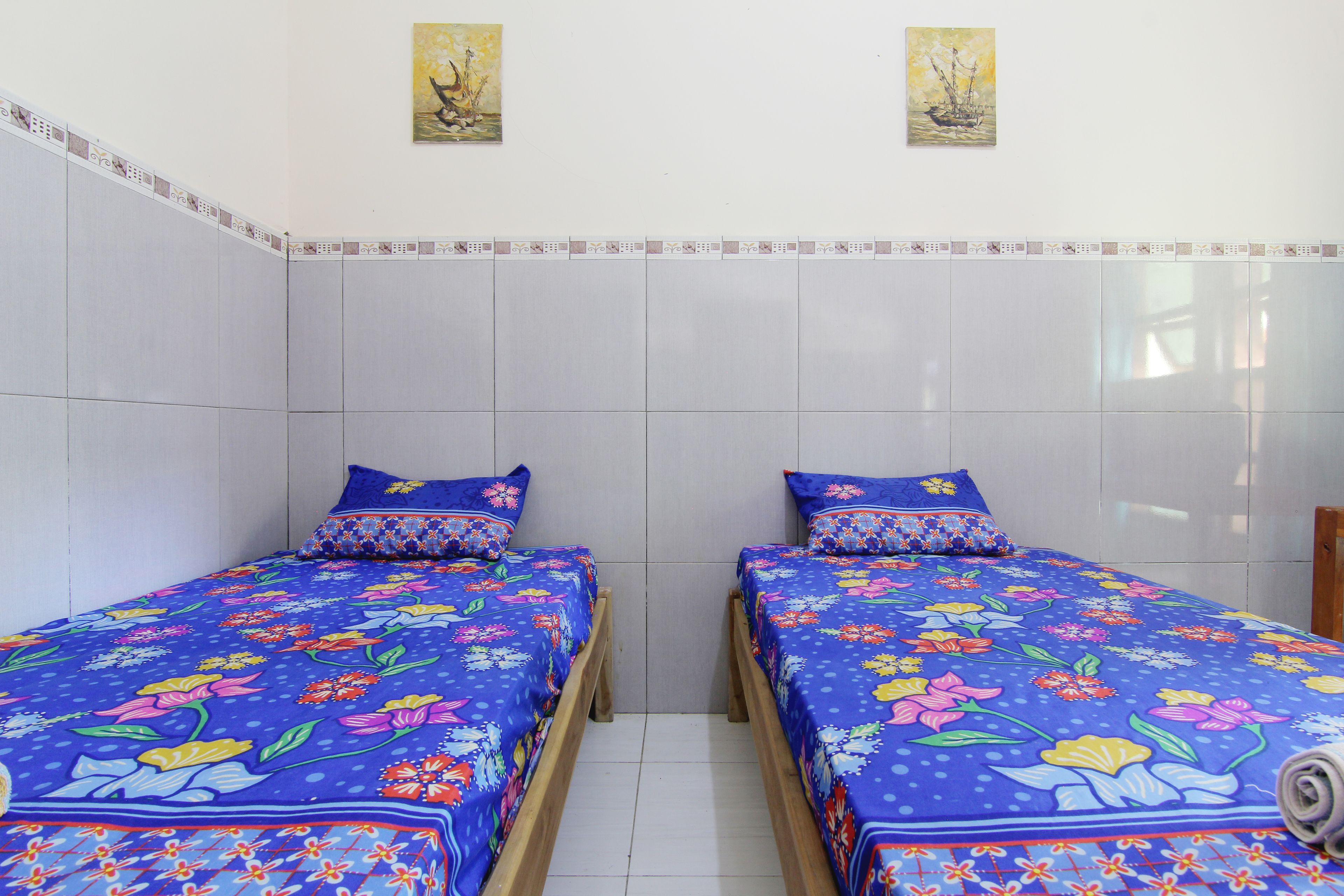 A room with two beds, each with a floral bedspread and matching pillows. There are two wall-mounted paintings above the beds.
