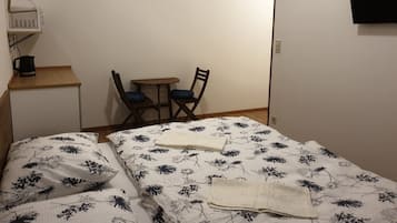 A small room with a bed, two chairs, a small table, a kettle, and a wall-mounted television.