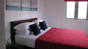 Una cama bien hecha con almohadas blancas y azul marino, una colcha roja y un cuadro con una playa sobre ella.
