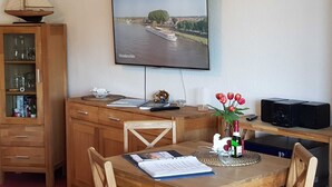 Une pièce comprenant un bureau en bois, un téléviseur fixé au mur qui diffuse une vue panoramique sur une rivière, une table à manger avec des chaises et un buffet contenant de la verrerie.