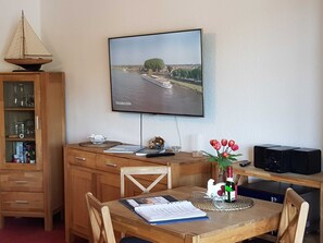 Una habitación con un escritorio de madera, un televisor montado en la pared que muestra una vista panorámica de un río, una mesa de comedor con sillas y un aparador con cristalería.
