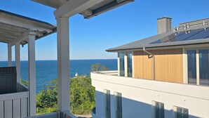 Un balcon avec vue sur la mer, un immeuble moderne avec un revêtement en bois et un ciel d’un bleu éclatant.