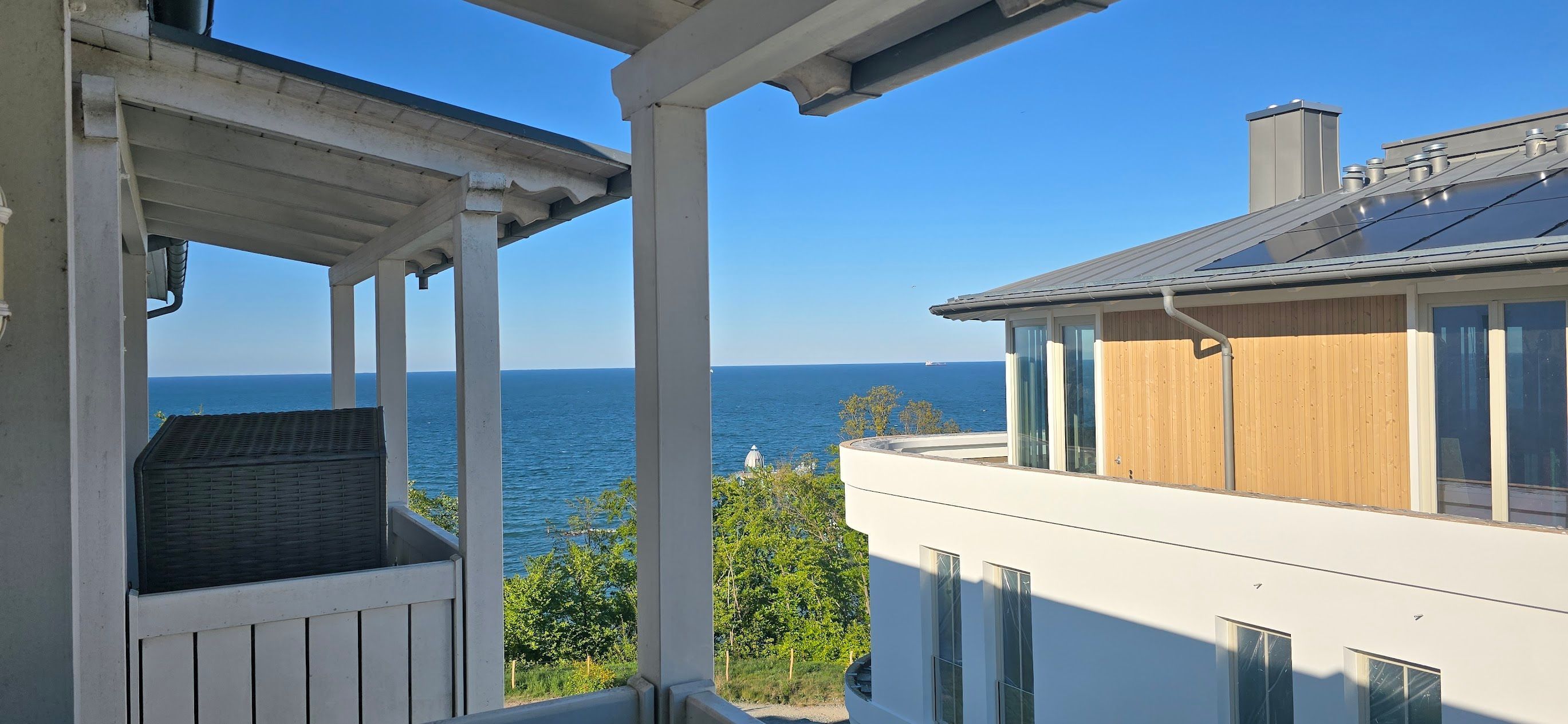 A balcony with a view of the sea, a modern building with wooden siding, and a clear blue sky.