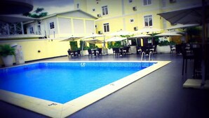 A swimming pool with lounge chairs and umbrellas, surrounded by a building with a yellow facade. - Royal Paragon Hotel (Port Harcourt)