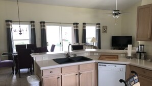 A modern kitchen with a dishwasher, a dining area with a table and chairs, a ceiling fan, and large windows with curtains. - Robin's Nest- Orlando Villa Vacation Rental. (Clermont)