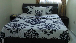 Un lit bien fait, recouvert d’une couvre-lit et de coussins aux motifs noir et blanc, une table de chevet avec une lampe, et une fenêtre avec des rideaux.
