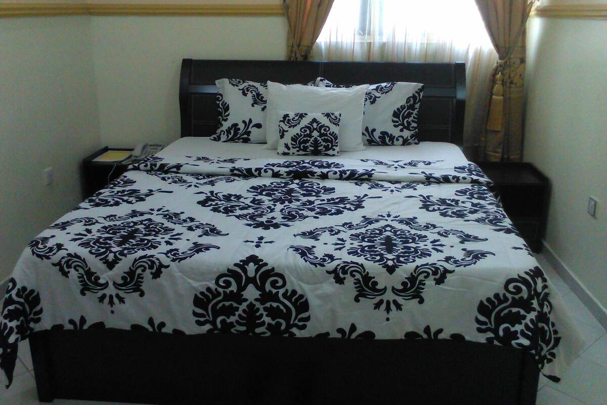 Una cama bien tendida con colcha y almohadas de estampado blanco y negro, una mesita de noche con una lámpara y una ventana con cortinas.