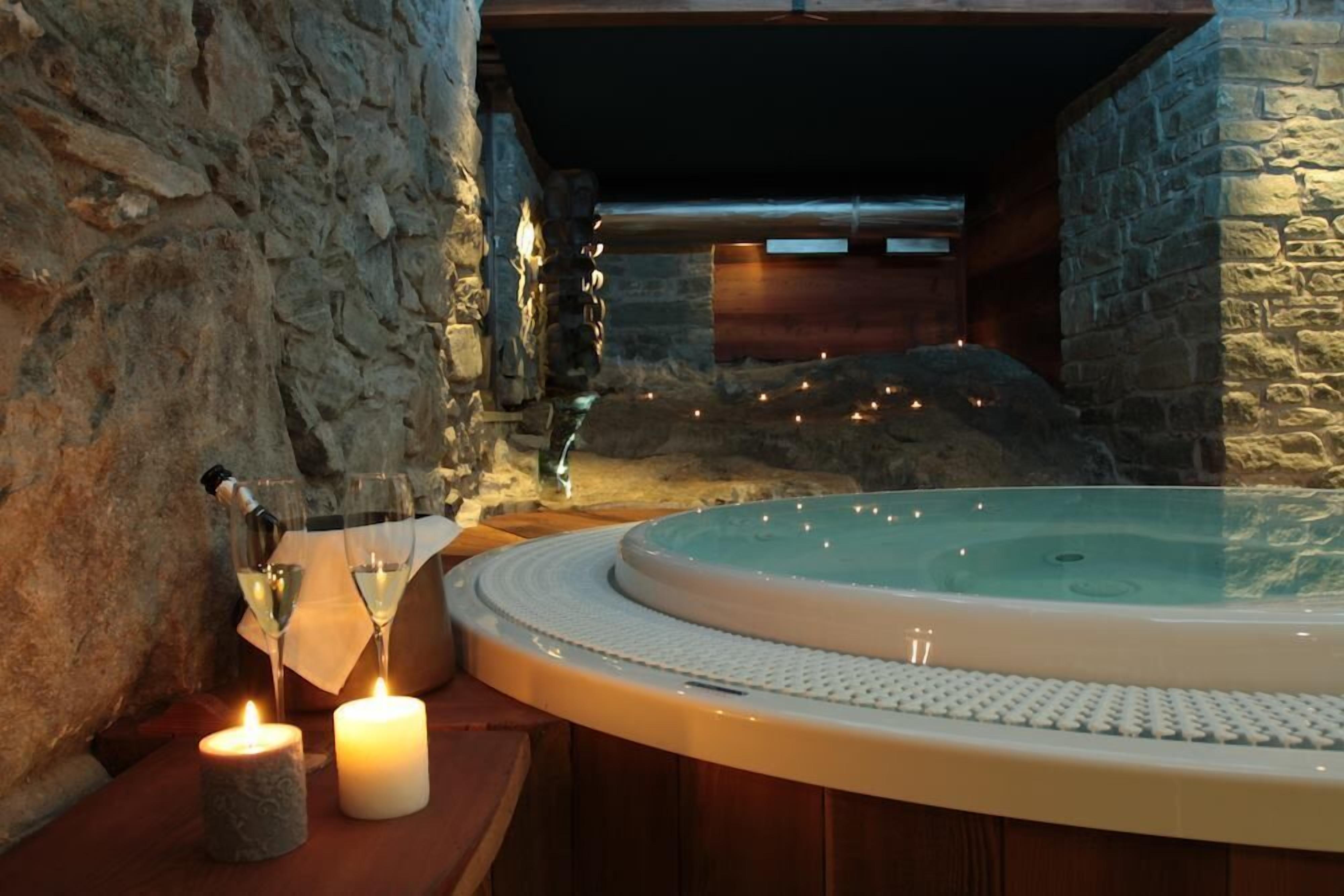 A stone-walled room with a hot tub, candles, and glasses of wine.