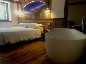 A bedroom with a wooden floor, a bed with white bedding, a large bathtub, and a wall-mounted light fixture. - Le Moulin des Aravis (Pontboset)