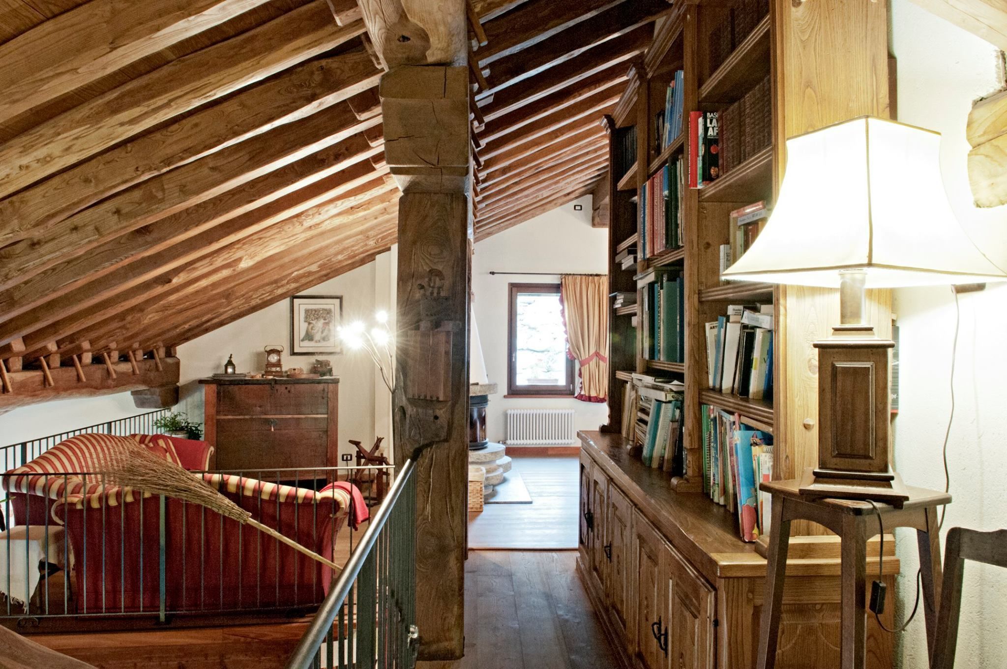 Amplia habitación en el ático con vigas de madera a la vista, un sofá rojo, un armario de madera lleno de libros y una lámpara.