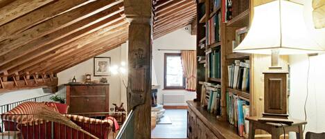 Amplia habitación en el ático con vigas de madera a la vista, un sofá rojo, un armario de madera lleno de libros y una lámpara.