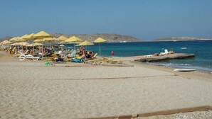 A beach with sun loungers and umbrellas, a pier extending into the sea, and rocky hills in the background. - Porto Paros (Paros)